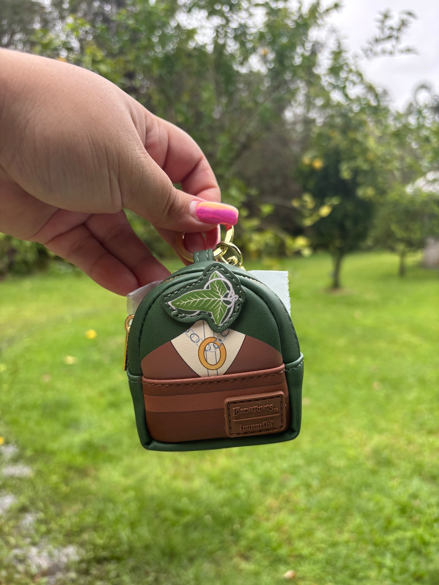 A hand with pink nail polish holds a small green and brown mini backpack keychain outdoors against a blurred green lawn and trees. The backpack features a green top, brown base, a white circular emblem, and a green leaf patch.