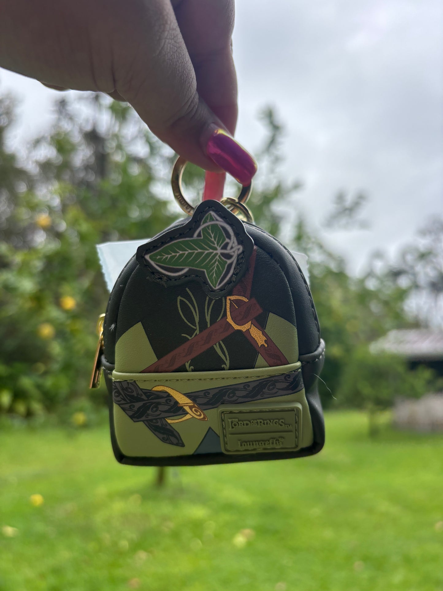 A hand with pink nail polish holds a small printed mini backpack keychain outdoors against a blurred green lawn and cloudy sky. The backpack-style keychain has a black, olive green, and tan camouflage-like print with gold-tone hardware.